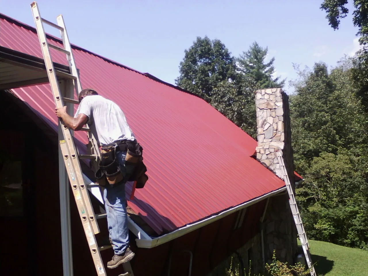 Roofer on a ladder beside a red metal roof and stone chimney — Soffit & Fascia Repair by Roofing Brothers in Flat Top